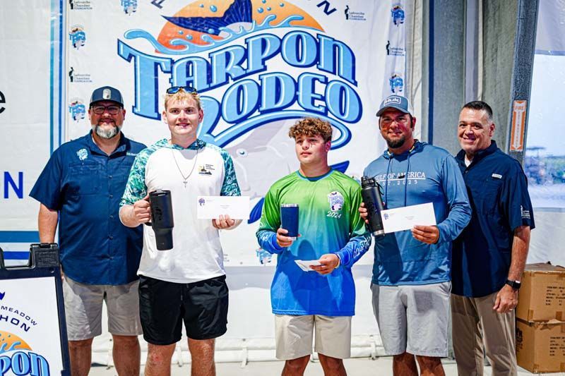 A group of men are posing for a picture in front of a tarpon rodeo banner.