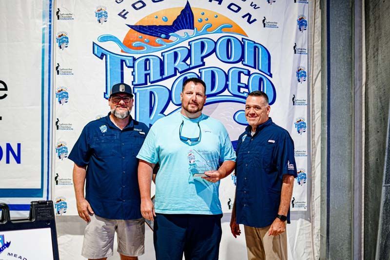 Three men are standing in front of a tarpon rodeo banner.