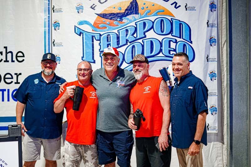 A group of men are posing for a picture in front of a tarpon rodeo banner.