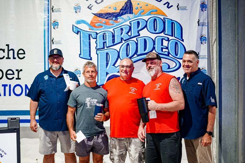 A group of men are posing for a picture in front of a tarpon rodeo banner.