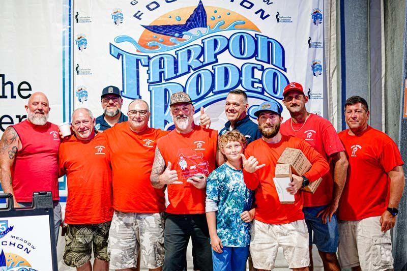 A group of men are posing for a picture in front of a tarpon rodeo banner.