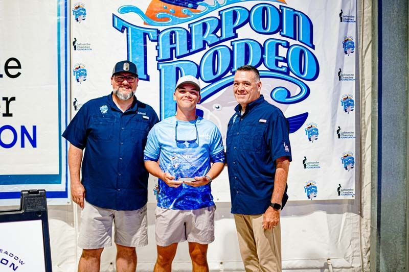 Three men are posing for a picture in front of a tarpon rodeo banner.