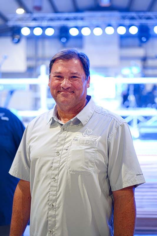 A man in a grey shirt is standing in a room and smiling.