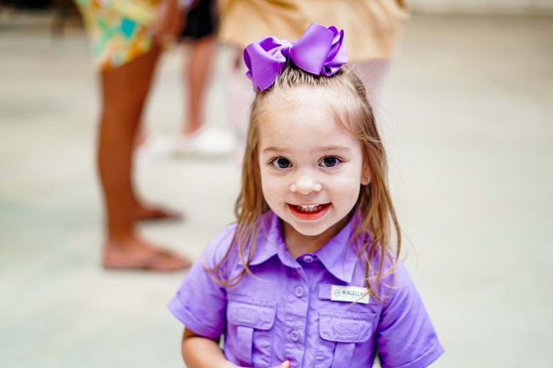 A little girl wearing a purple shirt and a purple bow is smiling for the camera.