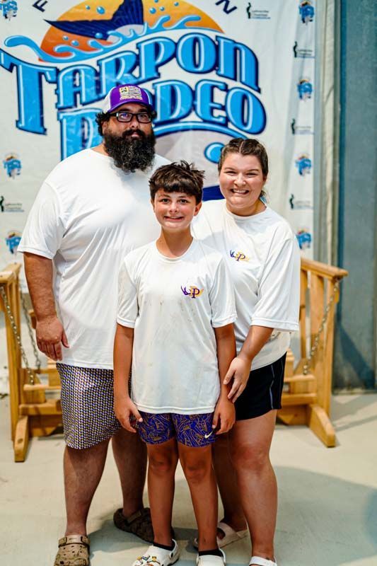 A man , woman and child are posing for a picture in front of a tarpon rodeo sign.