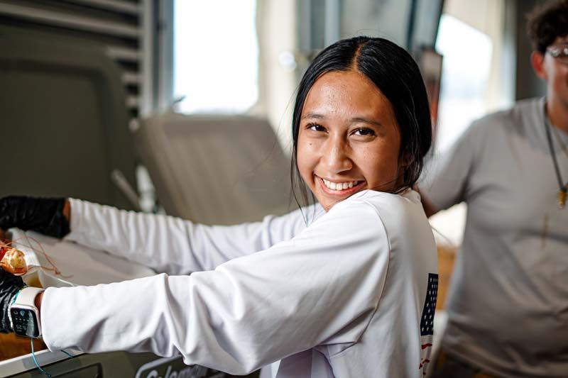 A woman in a white shirt is smiling while standing in a kitchen.