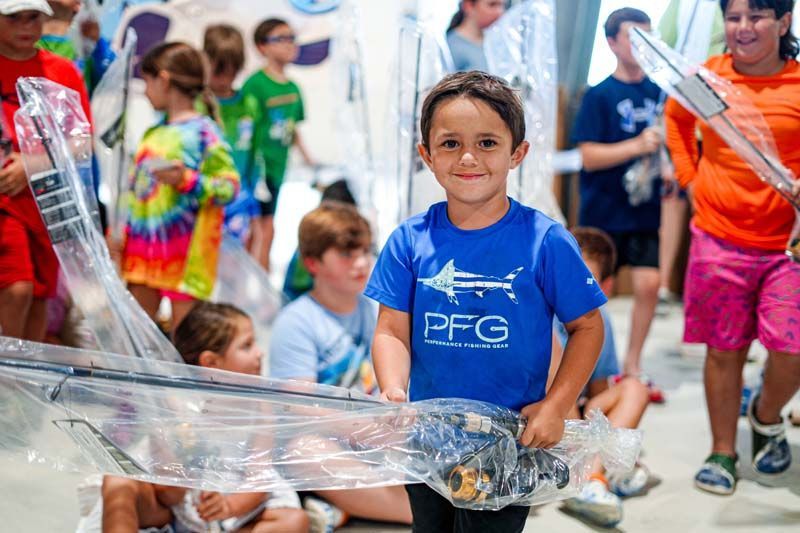 A young boy in a blue shirt is holding a plastic bag in front of a group of children.