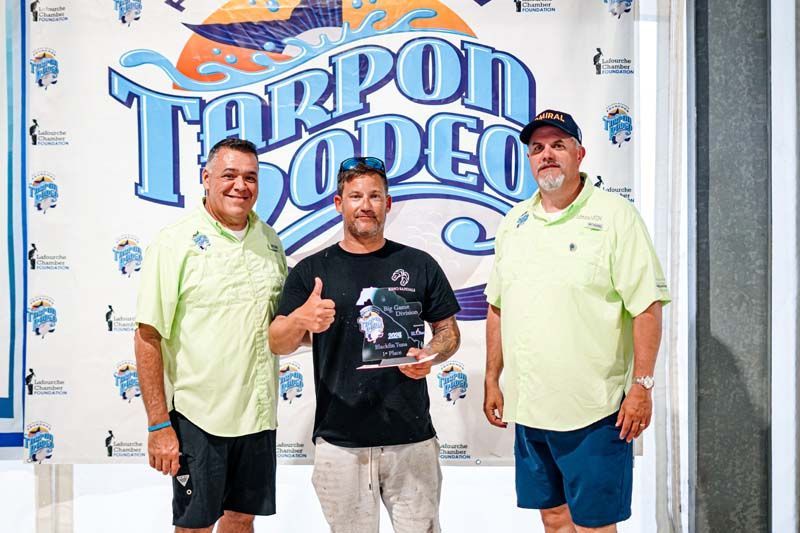 Three men are posing for a picture in front of a tarpon rodeo sign.