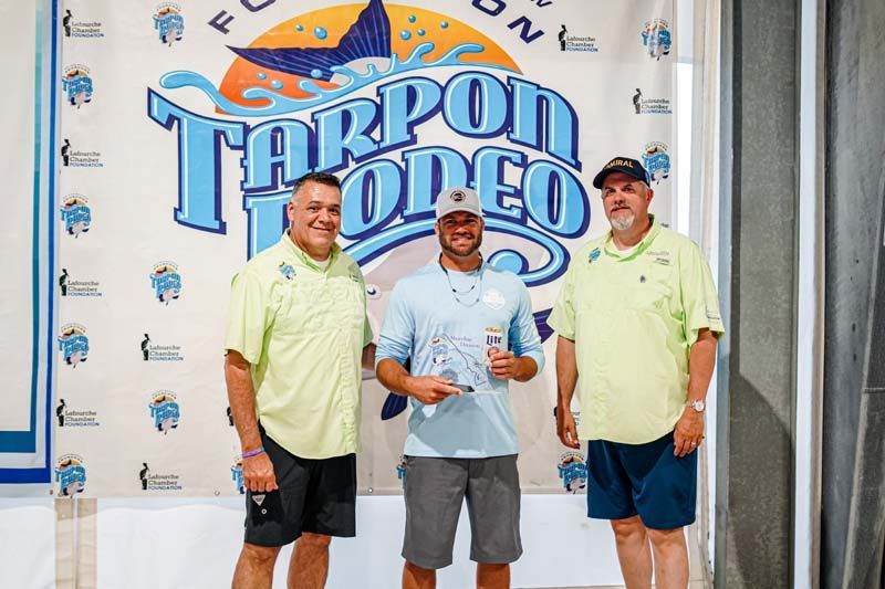 Three men are standing in front of a tarpon rodeo banner.