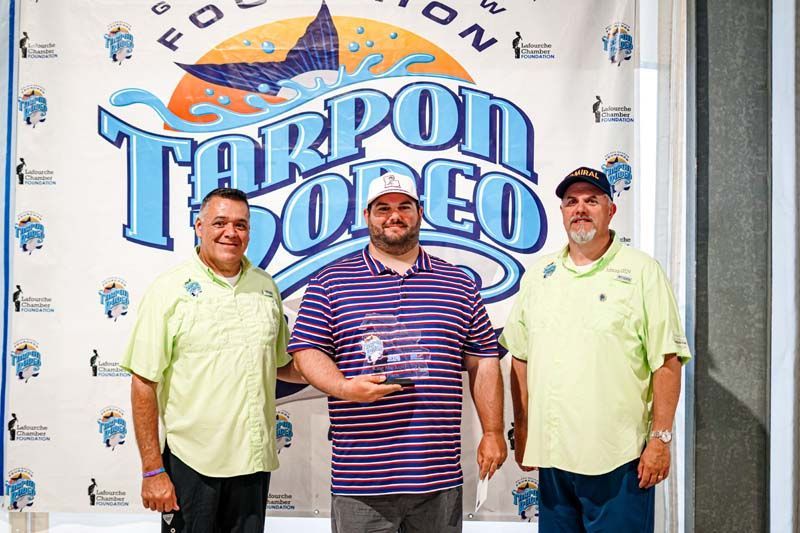 Three men are posing for a picture in front of a tarpon rodeo sign.