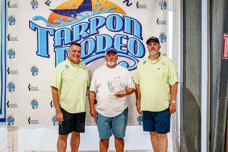 Three men are standing in front of a tarpon rodeo sign.