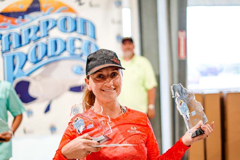 A woman in a red shirt is holding a trophy in front of a sign.