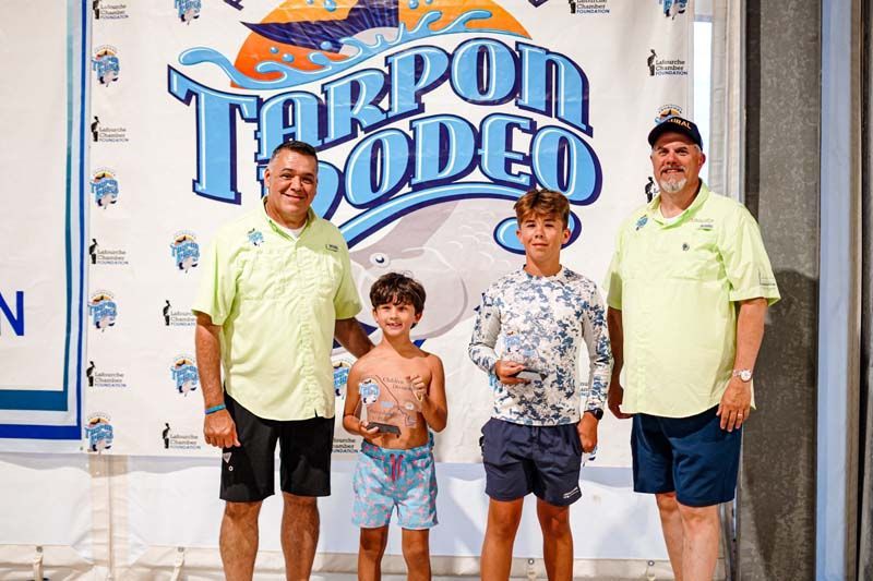 A group of men and two young boys are standing in front of a tarpon rodeo banner.