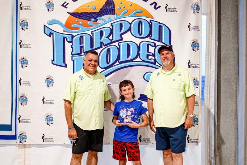 Two men and a boy are standing in front of a tarpon rodeo banner.