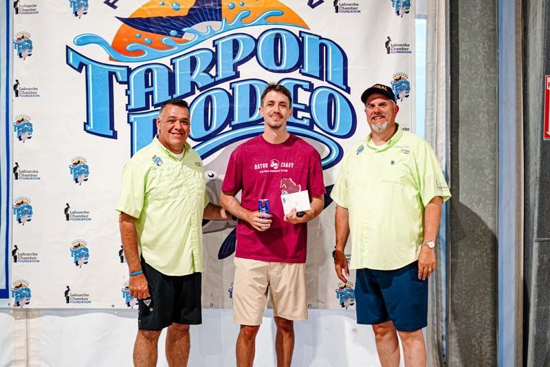 Three men are standing in front of a tarpon rodeo banner.