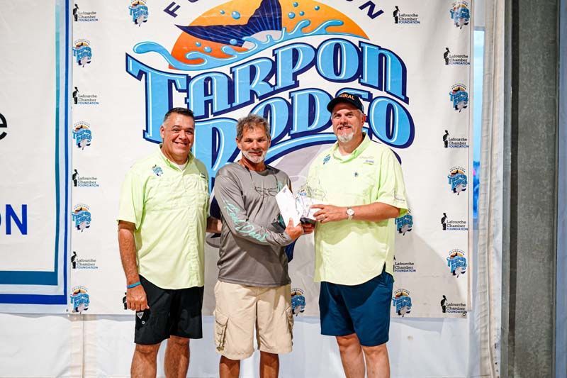 Three men are posing for a picture in front of a tarpon doo sign.
