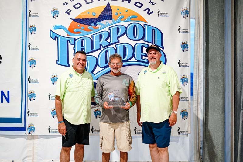 Three men are posing for a picture in front of a tarpon rodeo banner.