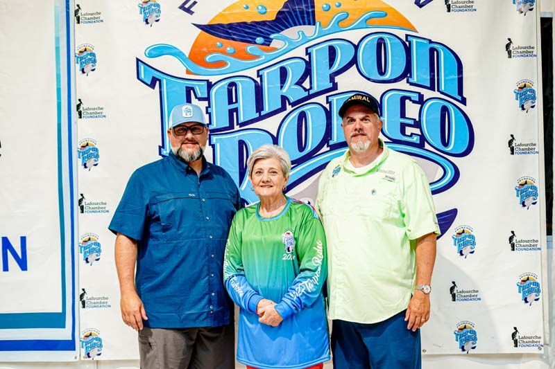 Three people are posing for a picture in front of a tarpon rodeo banner.
