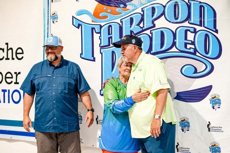 Two men are hugging a woman in front of a tarpon rodeo sign.