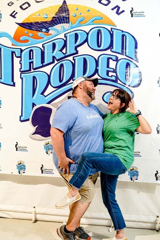 A man is holding a woman in his arms in front of a tarpon rodeo sign.