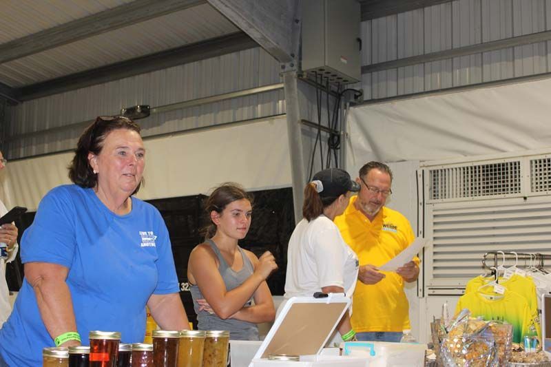 A woman in a blue shirt is standing in front of a table with jars of jam.