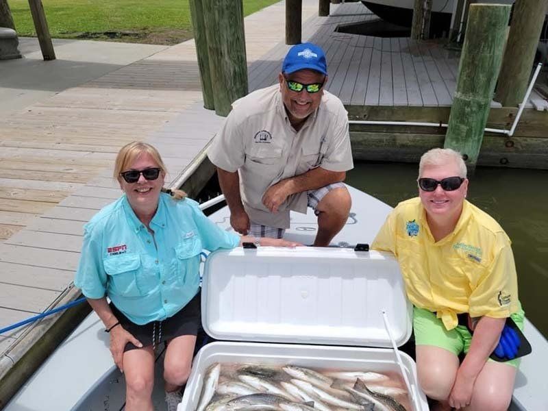 Three people are sitting on a boat with a cooler full of fish.