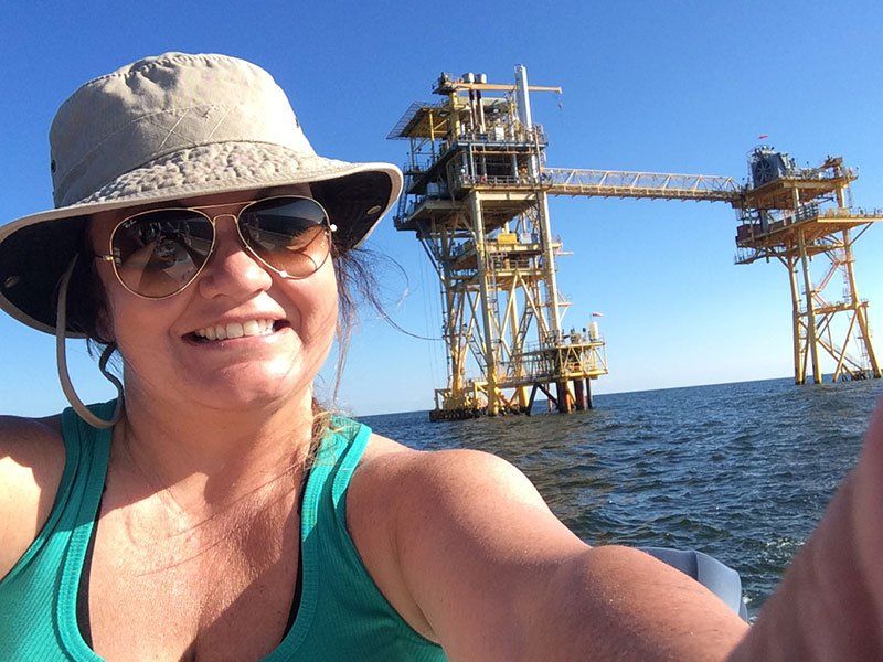 A woman wearing a hat and sunglasses is taking a selfie in front of an oil rig in the ocean.