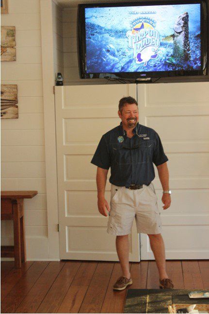 A man stands in front of a flat screen tv which shows a fishing show