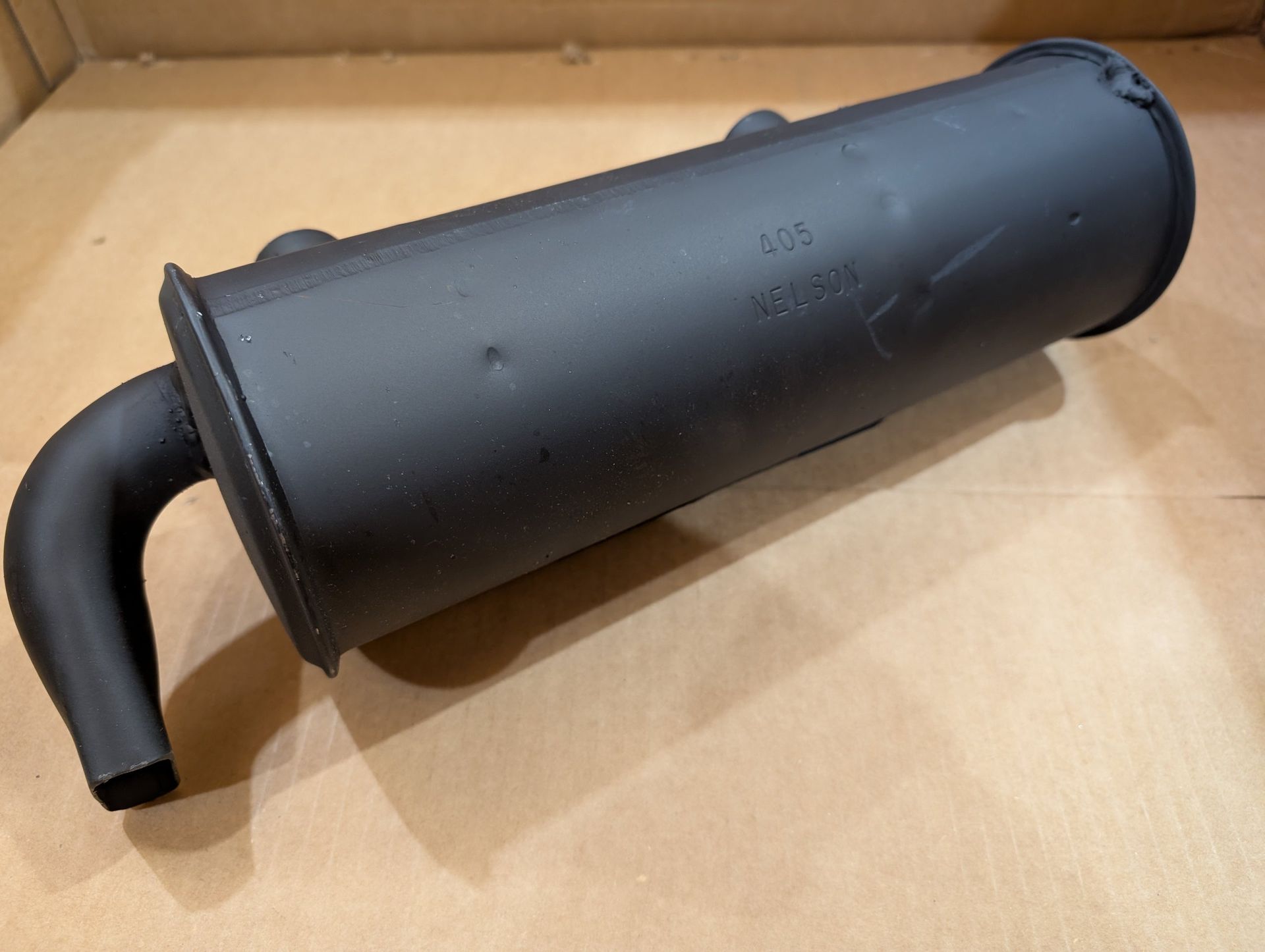 Black cylindrical muffler with a curved pipe, resting in a cardboard box.