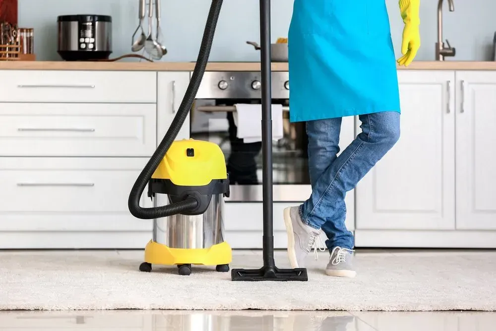 Person in Jeans and Blue Apron, Vacuuming a White Kitchen Floor — Jamac Cleaning Services in Merewether, NSW