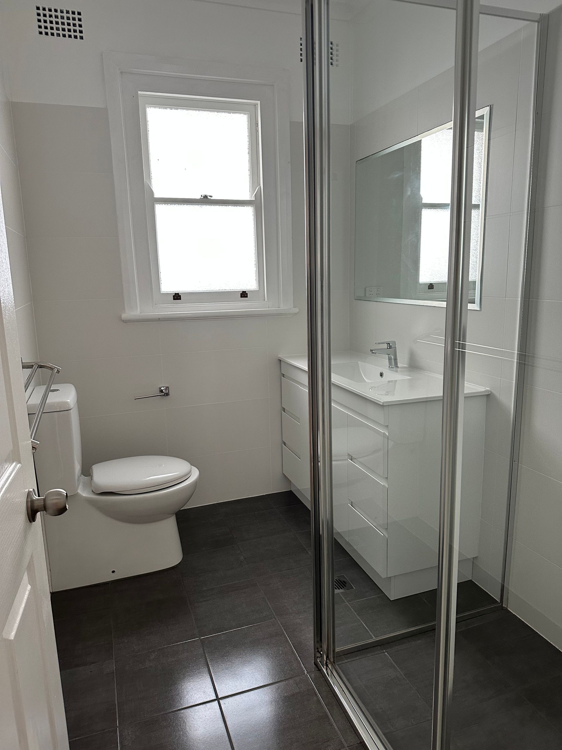 Bright bathroom with toilet, window, sink vanity, and glass shower enclosure, viewed from the doorway