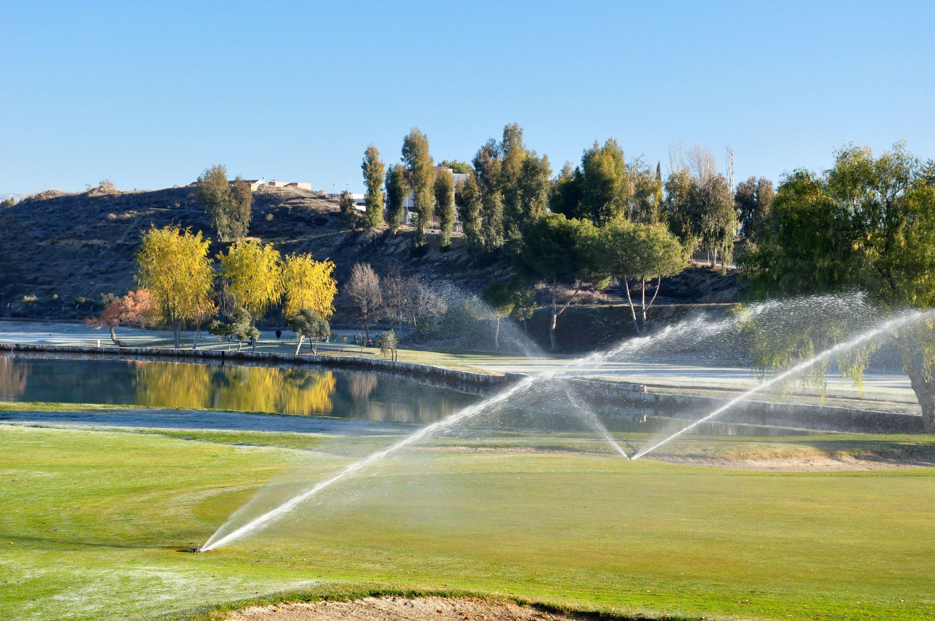 A sprinkler waters a green golf course lawn near a calm pond with golden autumn trees in the background.