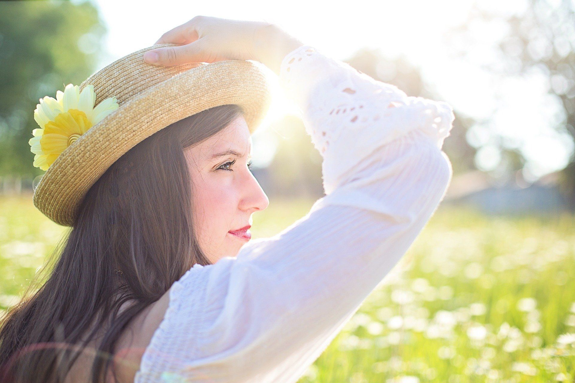 A woman wearing a straw hat with a yellow flower on it is sitting in a field of daisies.