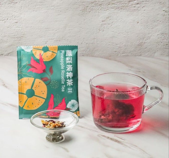 一包鳳梨洛神花茶、一杯泡好的紅茶和一小碟散裝茶葉，擺在大理石檯面上。