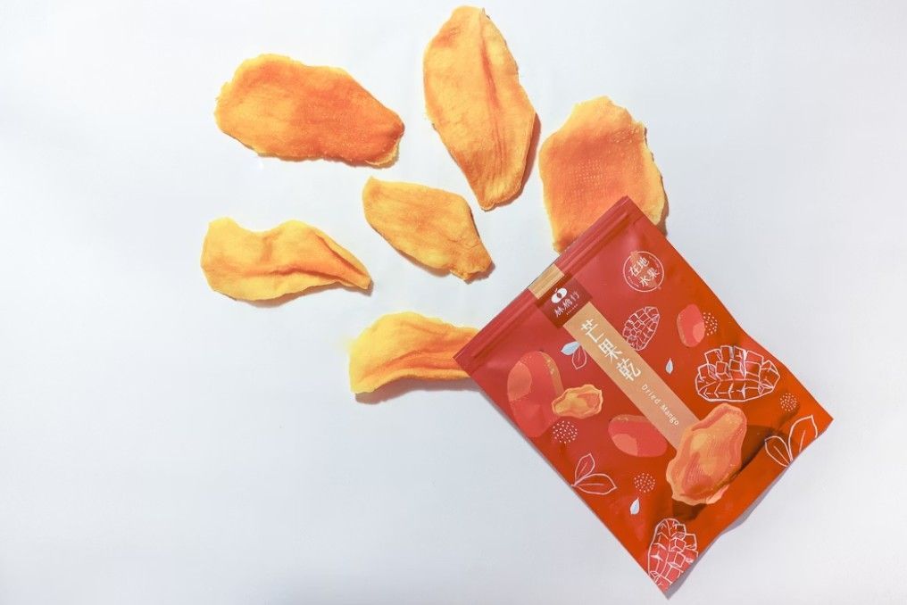 白色背景上散落著幾片芒果乾，旁邊是一個打開的橙色零食袋。