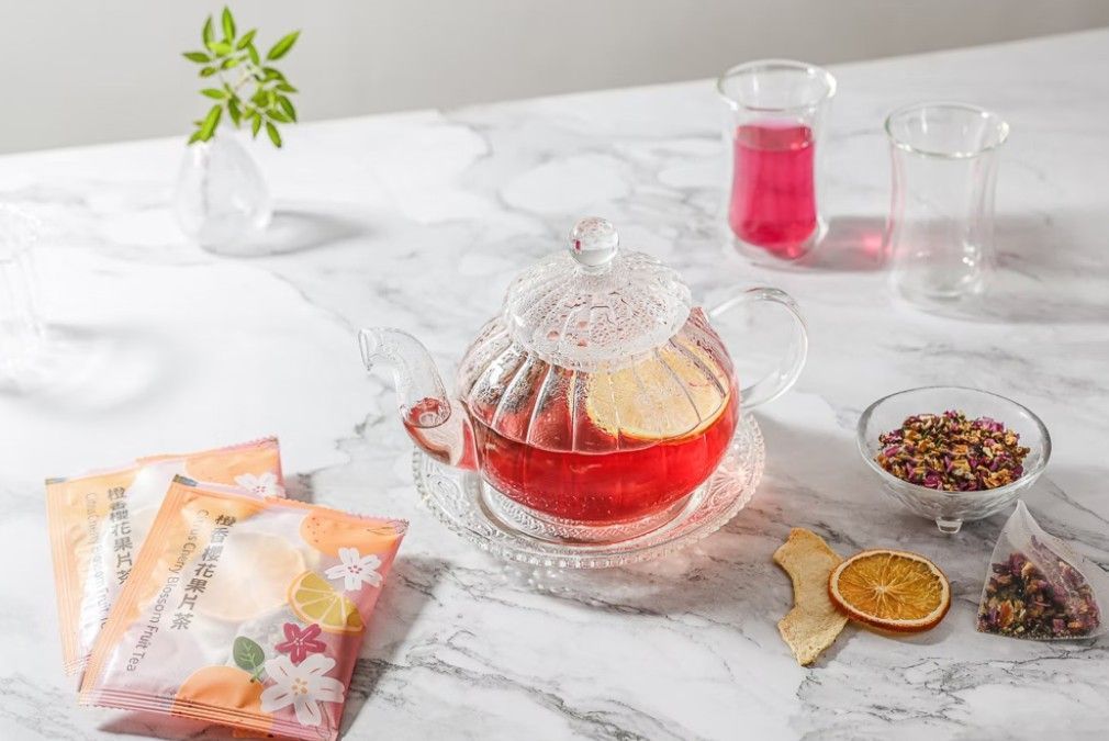 大理石桌上放著一個盛著鮮紅色花草茶的玻璃茶壺，旁邊還有一片柑橘、茶包、乾貨和玻璃杯。