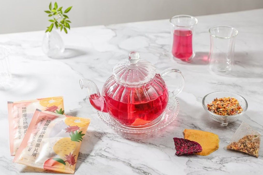 一個裝滿鮮紅色茶葉的玻璃茶壺放在大理石桌上，旁邊還有兩包茶葉、散裝茶葉和乾果片。