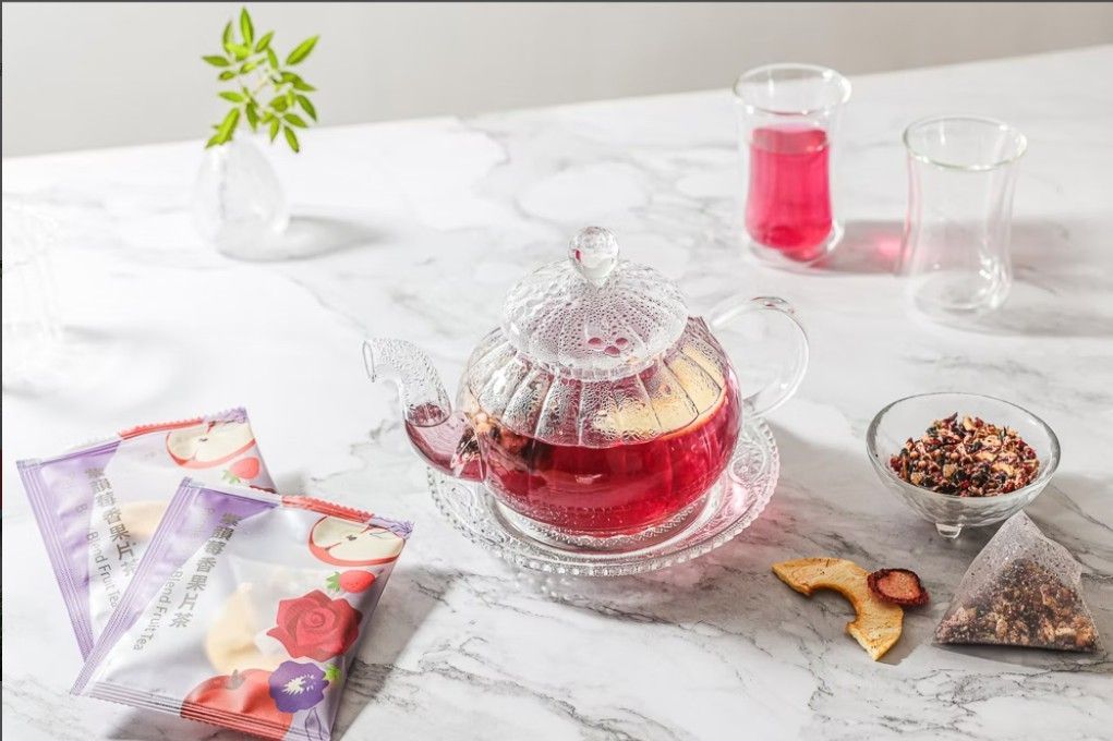 一個水晶茶壺裡裝滿了深紅色的水果茶，茶包、乾果和玻璃器皿放在大理石桌面上。