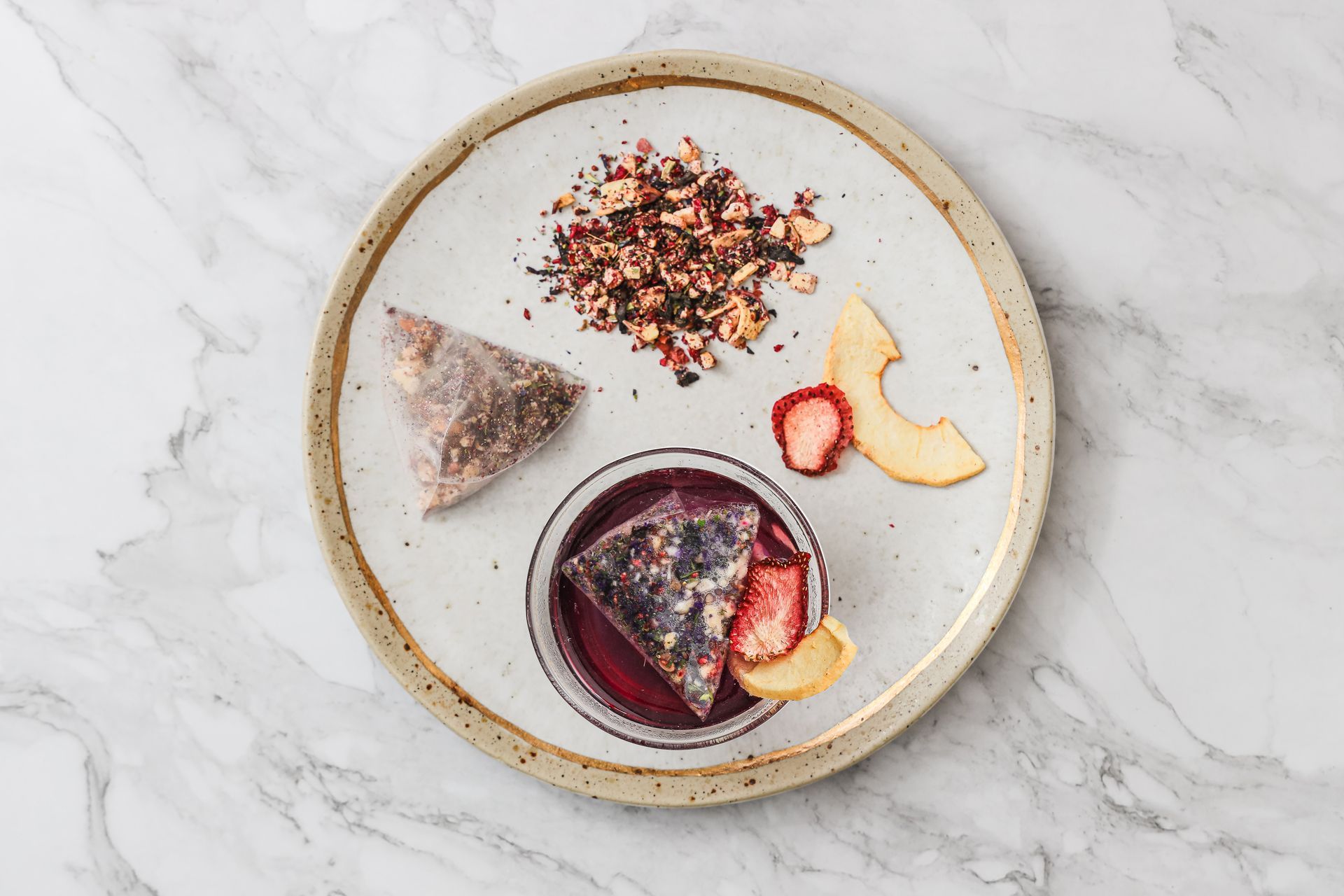 一個水晶茶壺裡裝滿了深紅色的水果茶，茶包、乾果和玻璃器皿放在大理石桌面上。