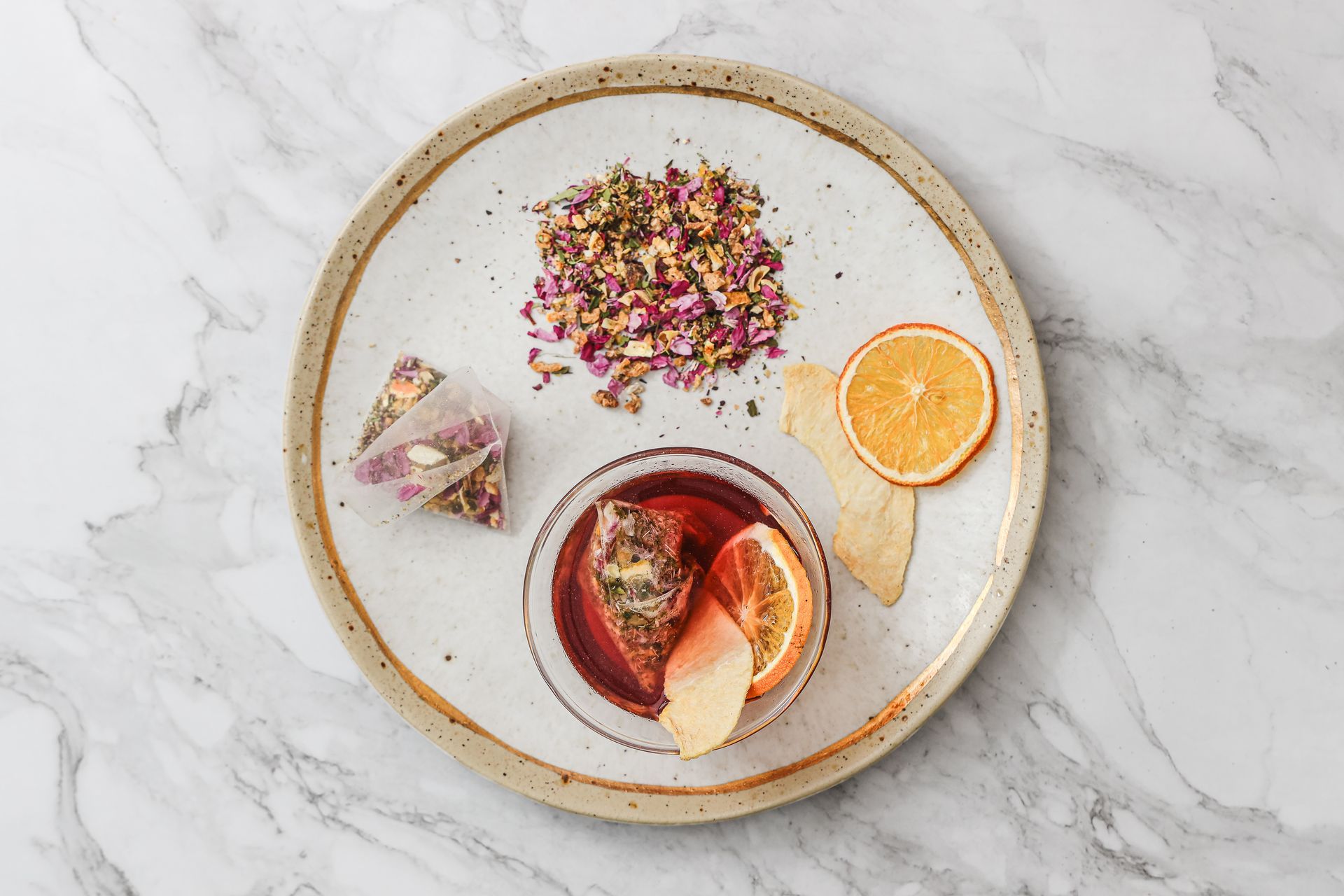 大理石桌上放著一個盛著鮮紅色花草茶的玻璃茶壺，旁邊還有一片柑橘、茶包、乾貨和玻璃杯。