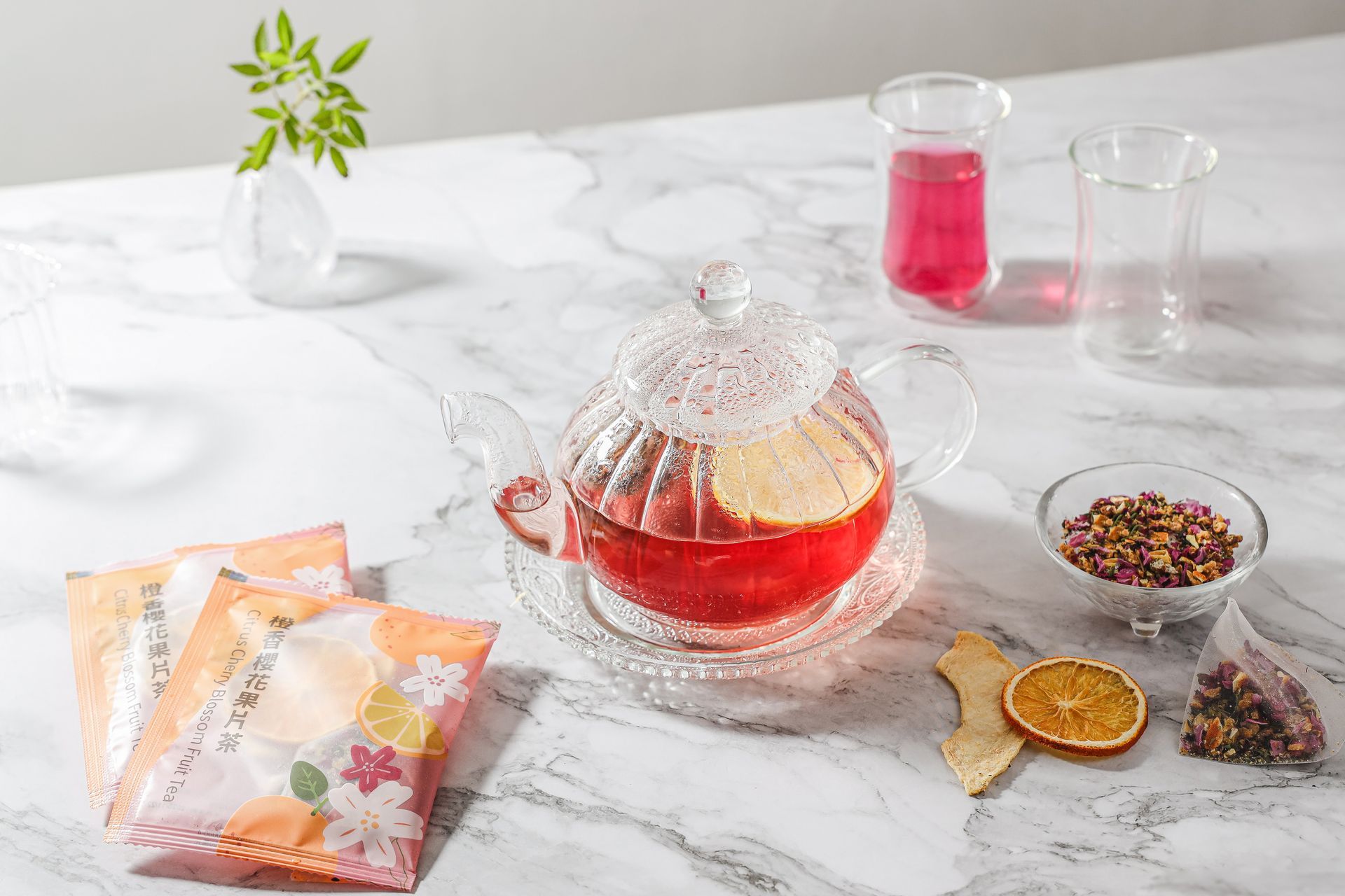 一個裝滿紅茶的玻璃茶壺放在大理石檯面上,周圍擺放著茶包、乾果、香草和一個玻璃杯。
