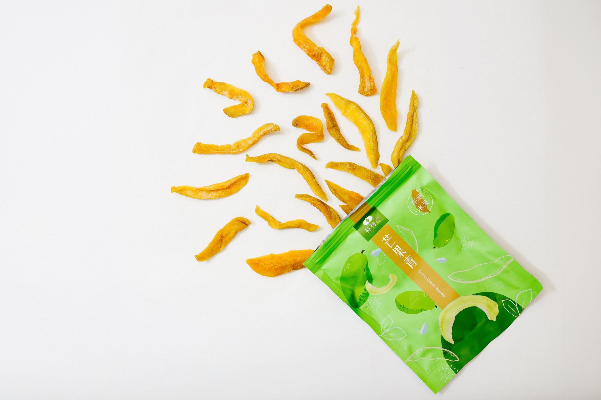 白色背景上的散落著芒果乾條，旁邊則是一個打開的綠色產品包裝袋。