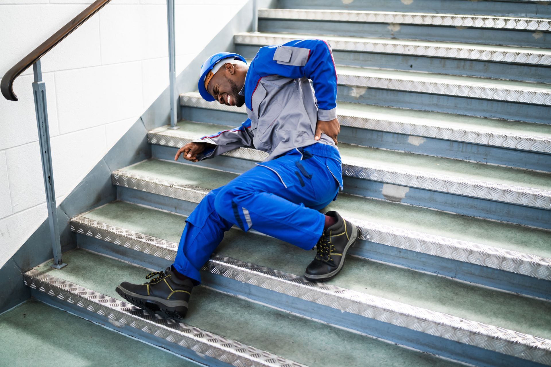 Person in blue work uniform sitting on metal stairs, clutching lower back.
