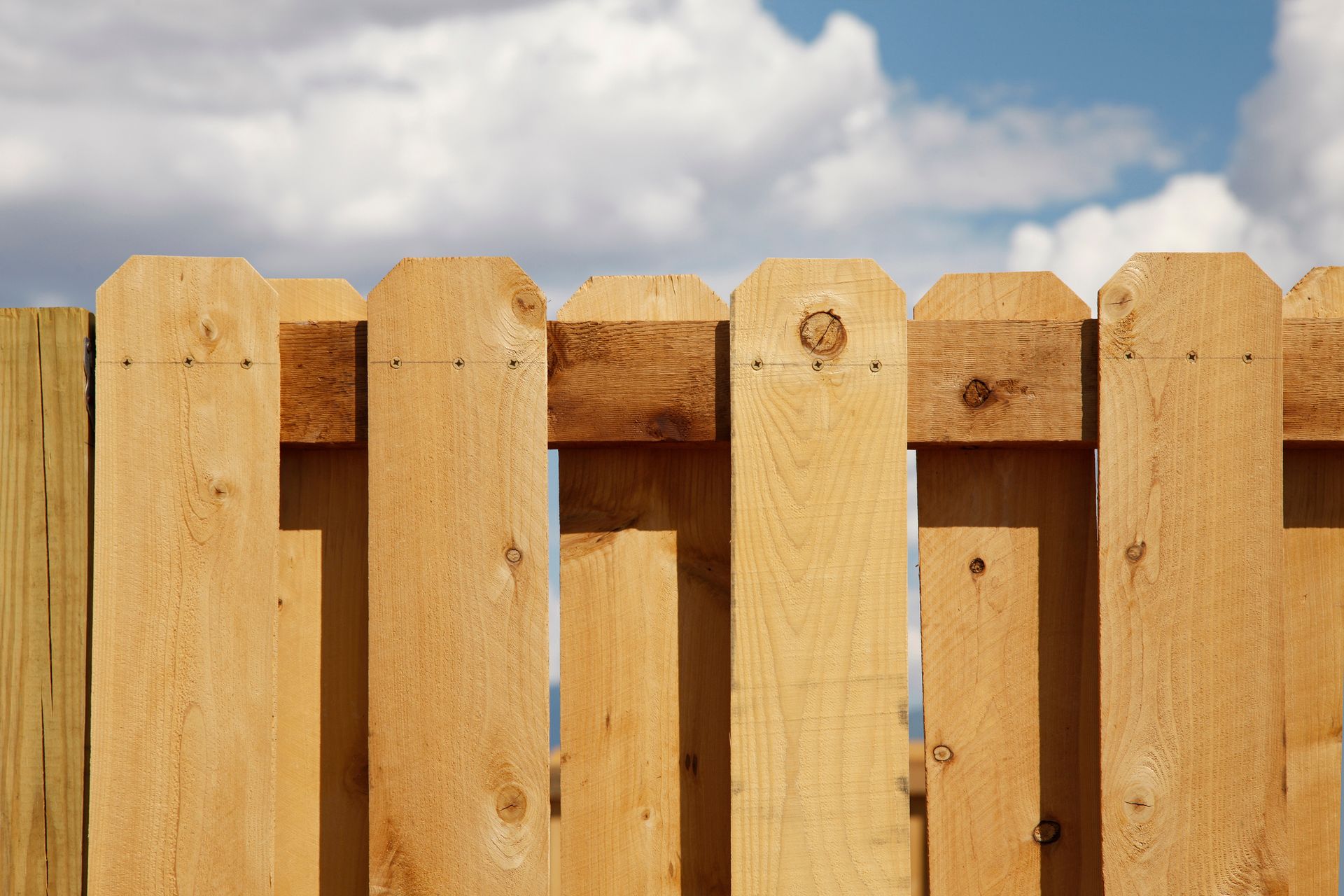 Close-up of cedar fence with angled tops and visible wood grain under a partly cloudy sky.