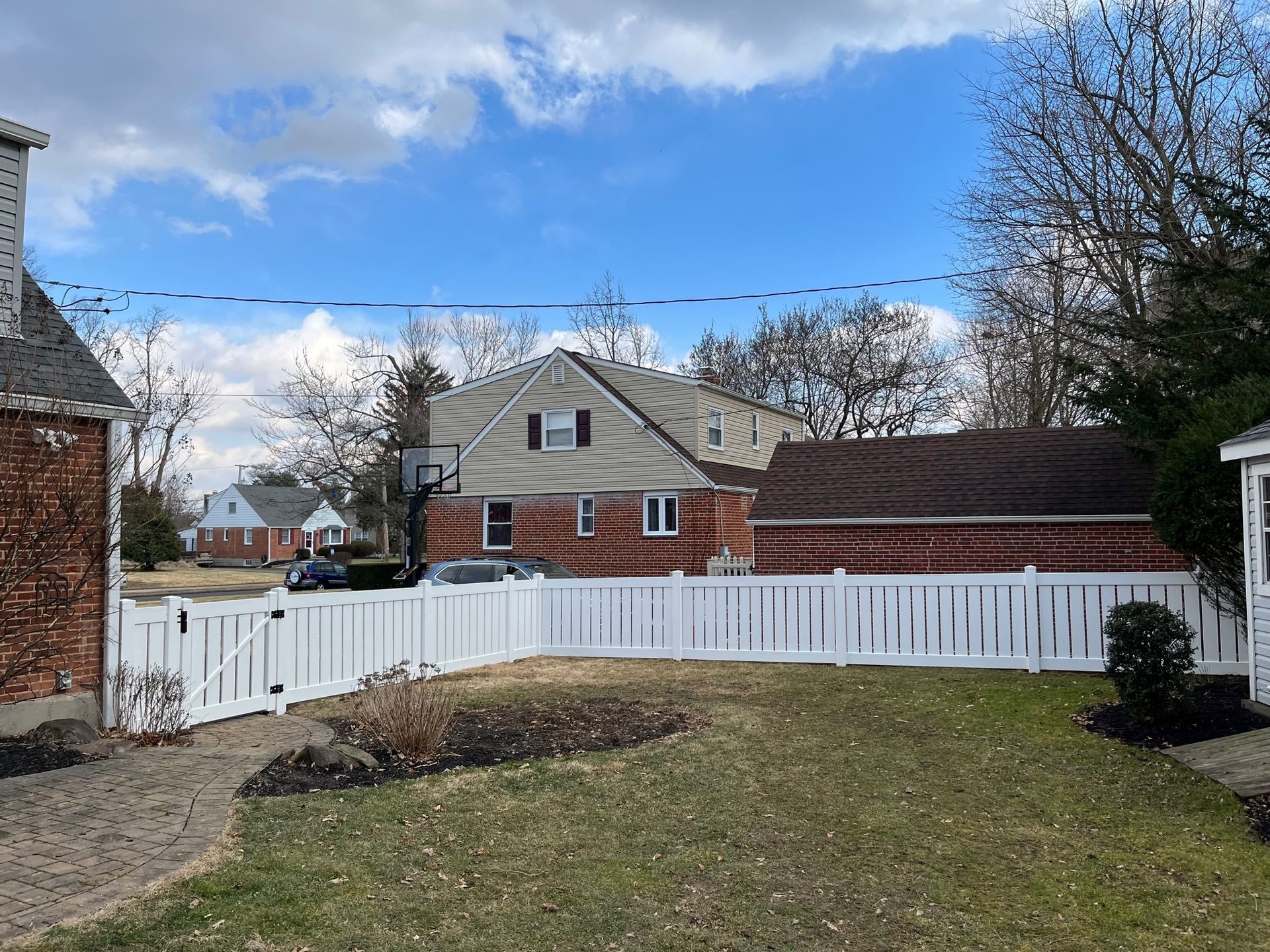 A white fence surrounds a brick house with a basketball hoop in the backyard.
