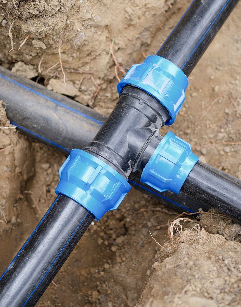 Black plastic pipes connected with a black T-fitting and blue connectors in a dirt trench.