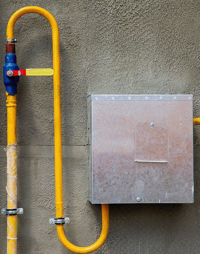 Yellow gas pipe with valve and metal box on a textured gray wall.