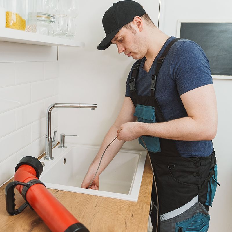 Plumber in blue shirt and overalls unclogging a white kitchen sink. A red plunger is on the counter.