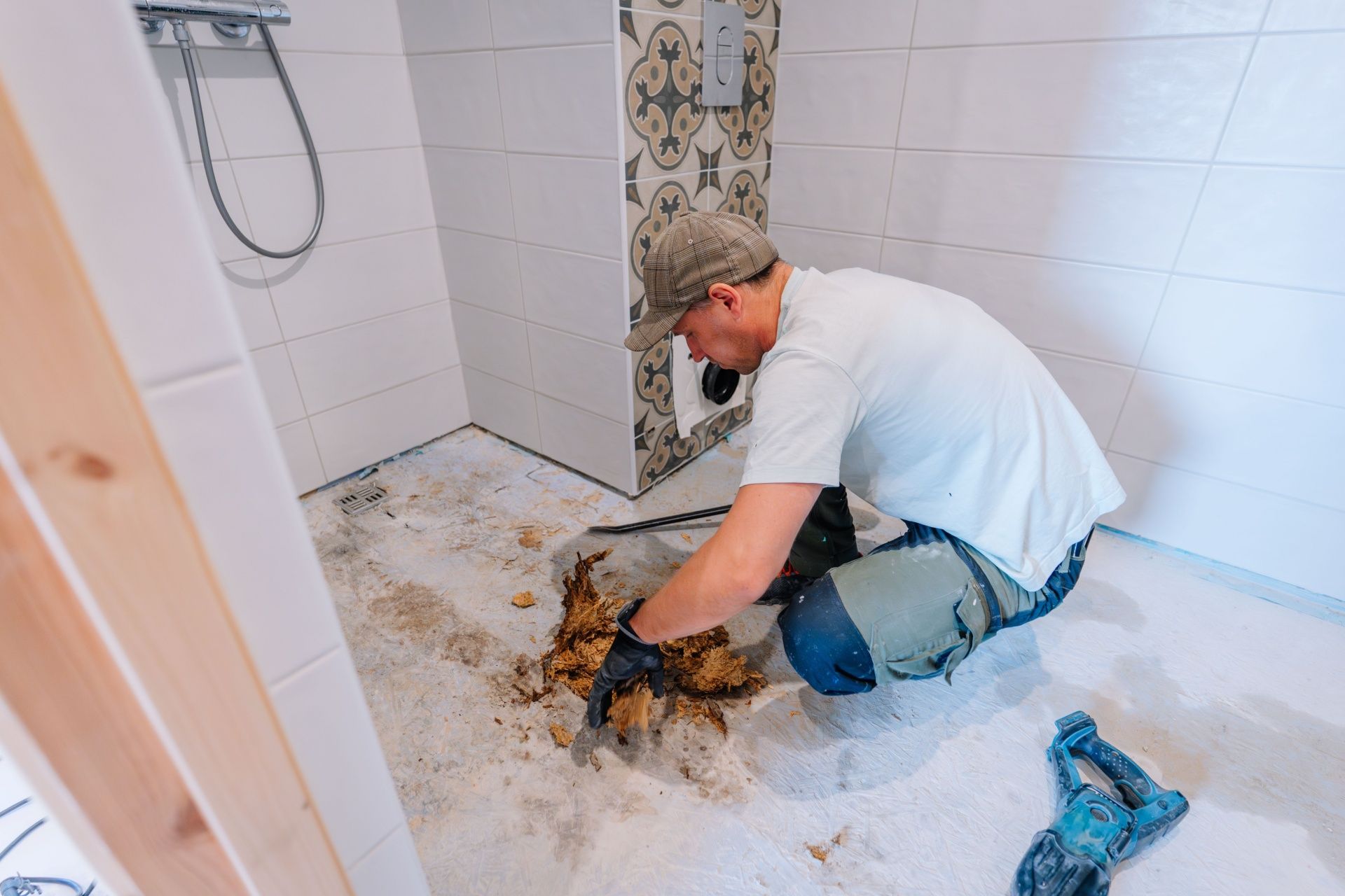 Person kneeling in bathroom removing debris, surrounded by white tile walls and shower fixtures.
