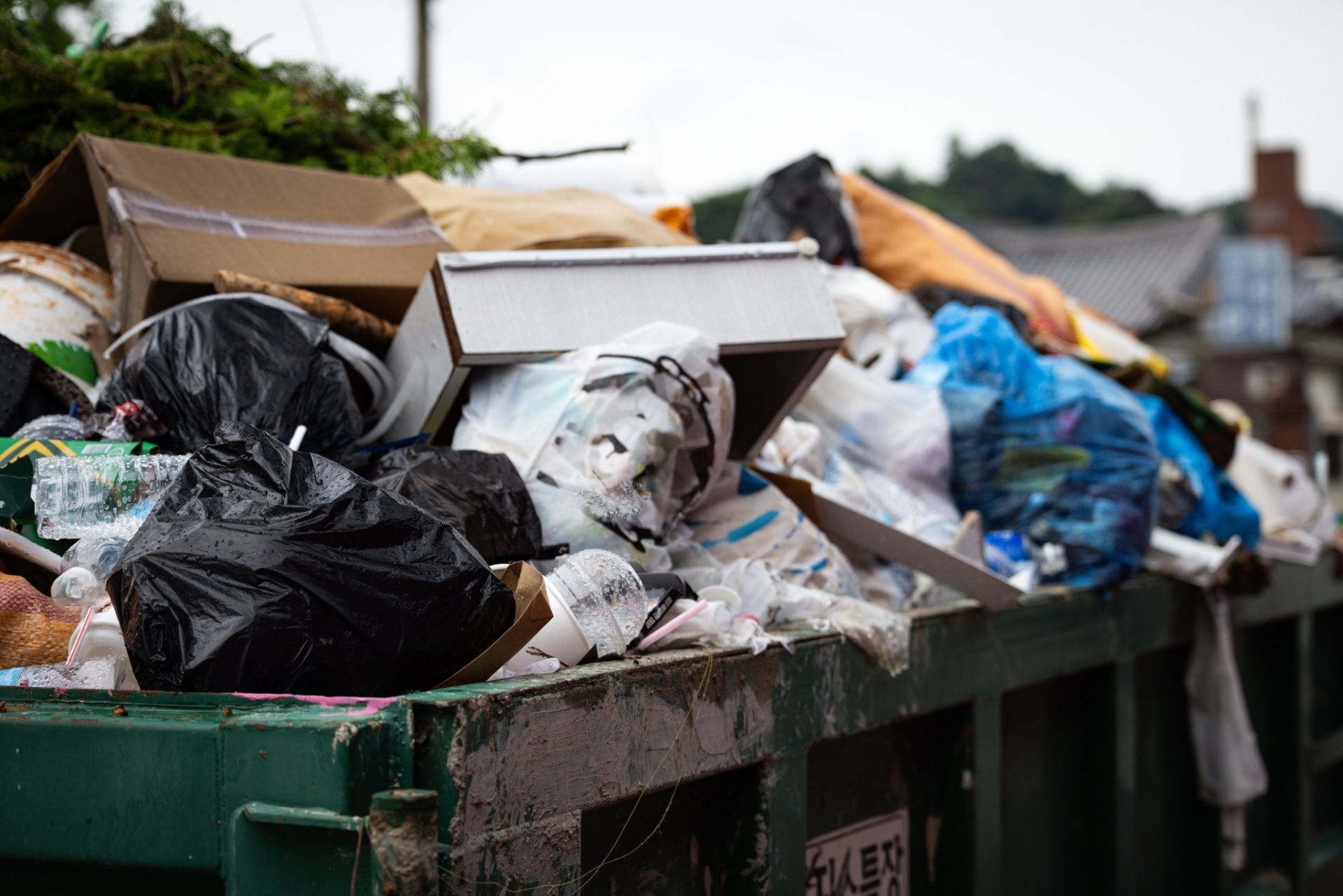Overflowing green dumpster filled with trash bags, boxes, and debris outdoors.